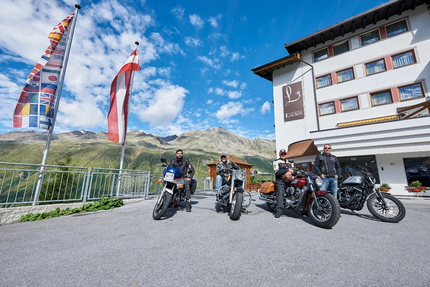 Bayrische Seenrunde Vier Motorradfahrer vor Bergkulisse und Hotelgebäude mit Flaggen im Wind
