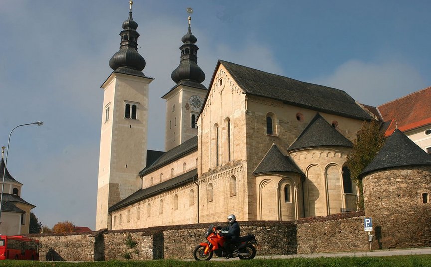 Person fährt Motorrad vor Klosterkirche mit doppelten Türmen bei klarem Himmel