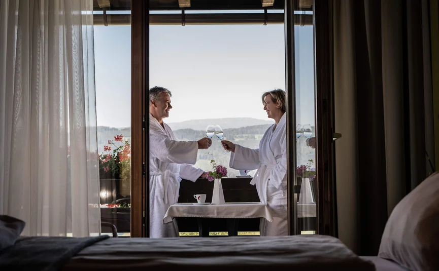 Couple in bathrobes toasting with glasses on balcony with mountain view