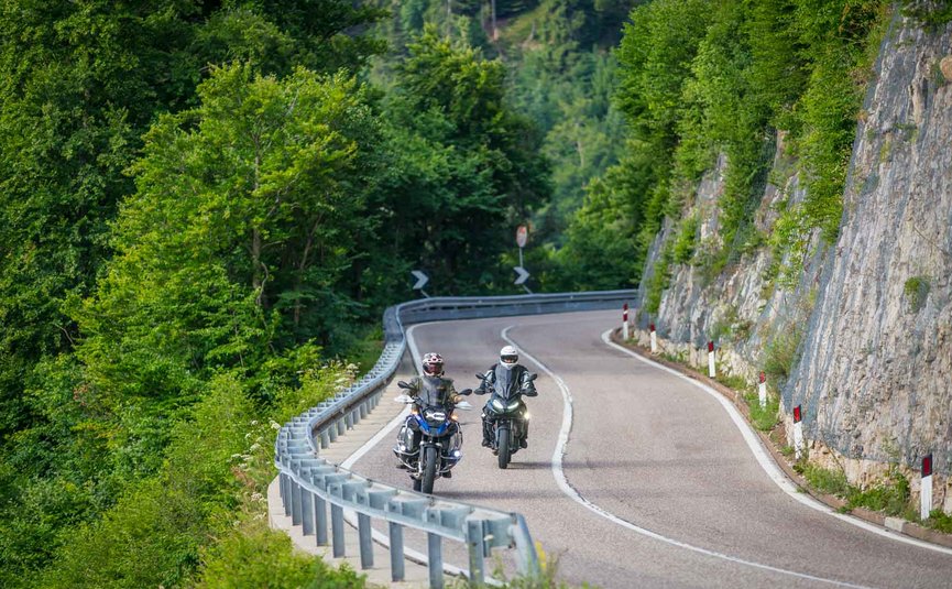 Zwei Motorradfahrer fahren auf kurviger Bergstraße mit Felsen und Bäumen