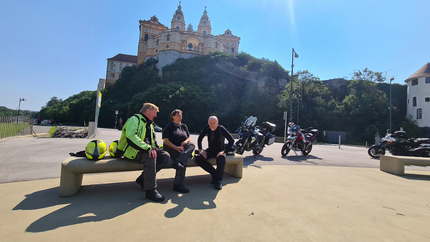 Wachautour Drei Motorradfahrer sitzen auf einer Bank vor einem Berg mit Schloss im Sonnenschein