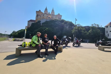 Three motorcyclists sitting on a bench near a hill with a castle on a sunny day