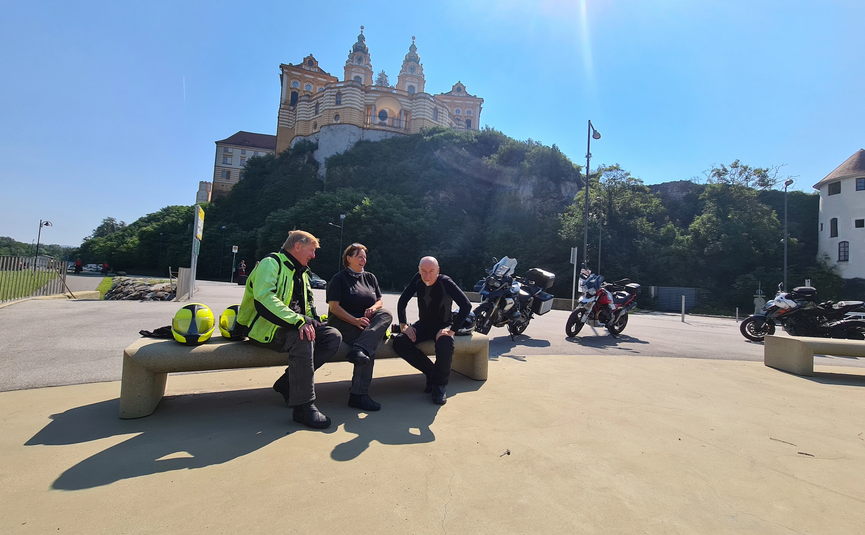Drei Motorradfahrer sitzen auf einer Bank vor einem Berg mit Schloss im Sonnenschein