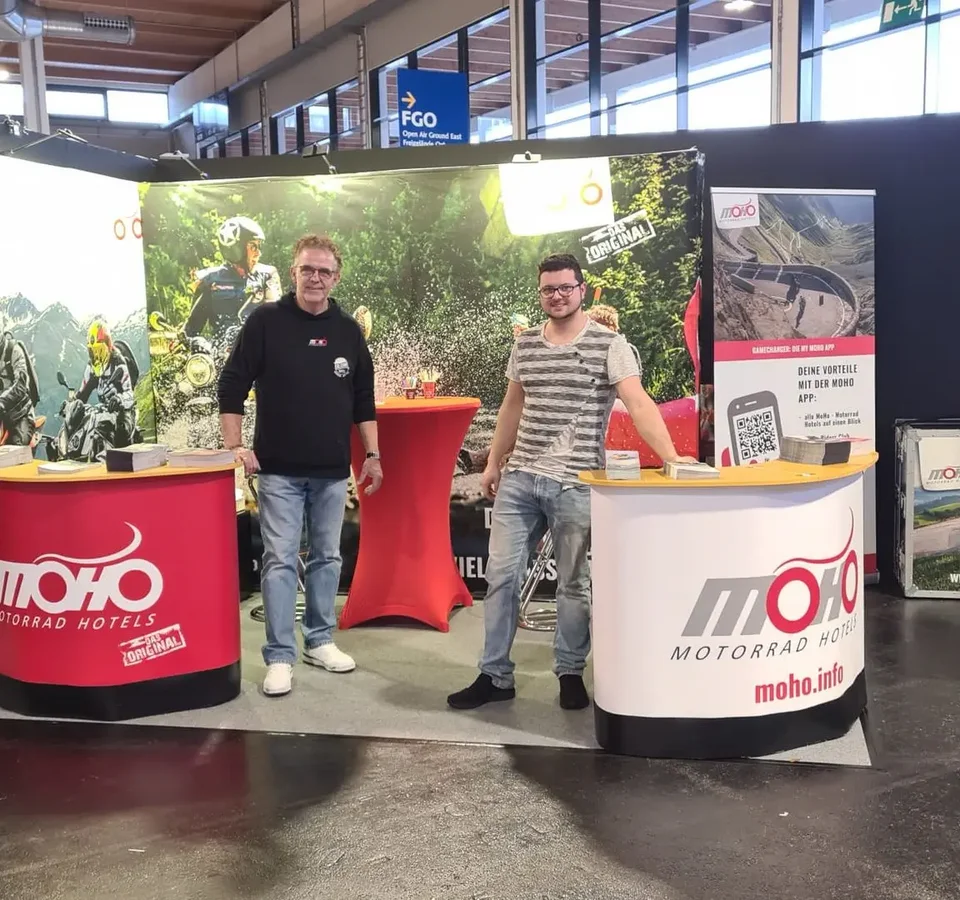 Motorradwelt Bodensee Zwei Männer am Messestand von MOHO Motorrad Hotels mit Mountainbike-Bildern