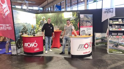 Zwei Männer am Messestand von MOHO Motorrad Hotels mit Mountainbike-Bildern