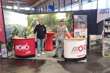 Zwei Männer am Messestand von MOHO Motorrad Hotels mit Mountainbike-Bildern