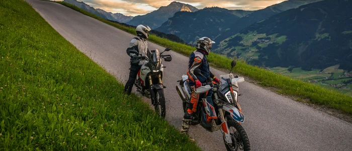 Zwei Motorradfahrer auf bergiger Straße mit Blick auf Tal und Berge bei Sonnenuntergang