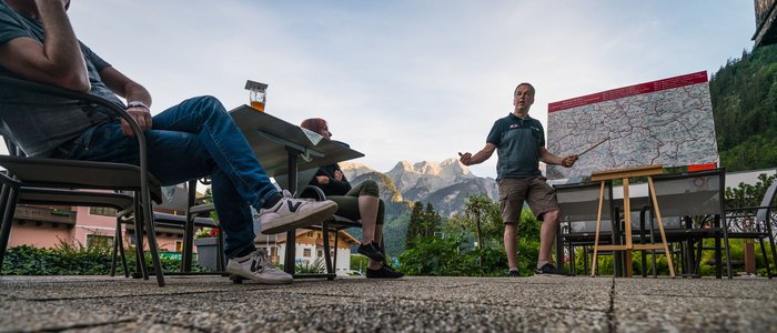 Mann erklärt Wanderkarte vor Bergkulisse, andere hören draußen zu