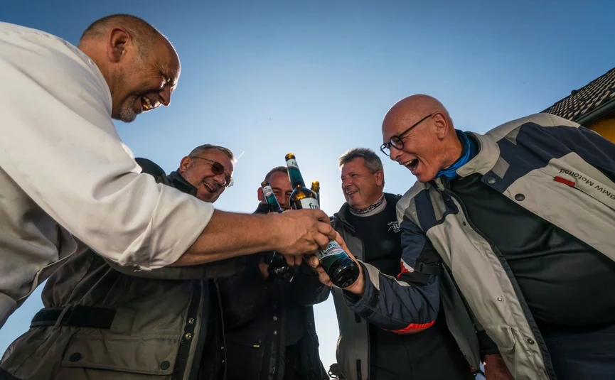 Five men laughing and clinking beer bottles under clear sky