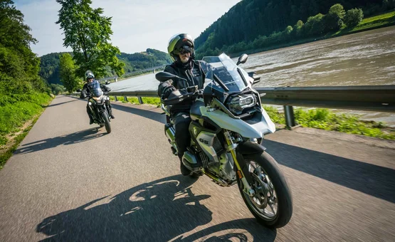 Two motorcyclists riding on a road beside a river in a mountainous landscape