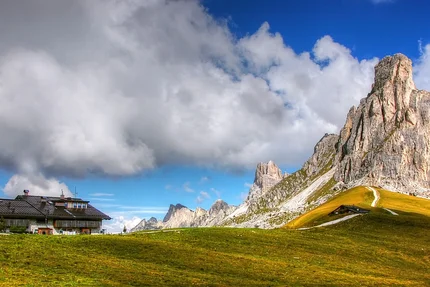 House by green hills and tall rocky peaks under a cloudy sky