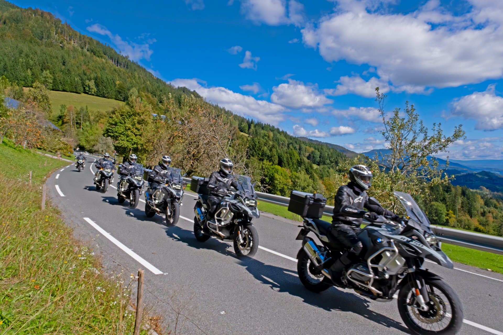 Motorradurlaub © Igor Rosina Gruppe Motorradfahrer auf Landstraße mit bewaldeten Hügeln und blauem Himmel