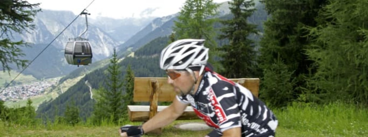 Mountainbikeurlaub Mountainbiker fährt bergauf auf Waldweg mit Seilbahn und Bergen im Hintergrund