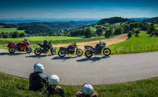 Drei Personen mit Helmen sitzen auf Wiese vor vier Motorrädern auf bergiger Landstraße