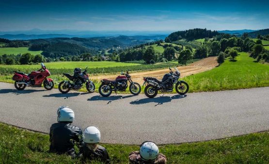 Three people with helmets sitting on grass near four motorcycles on a rural mountain road