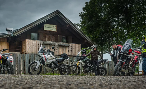 Motorradfahrer vor einer Holzhütte mit Motorrädern bei bewölktem Himmel