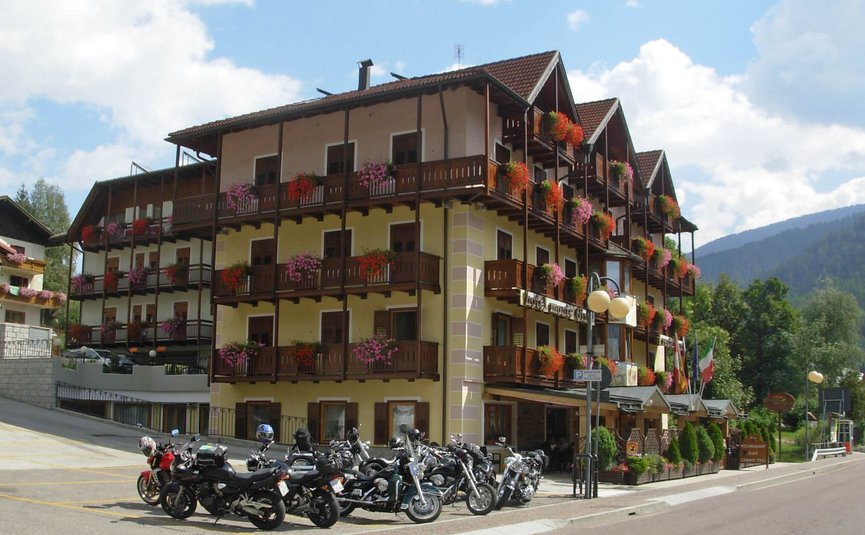 Hotel Monte Giner **** © Marisol s.n.c.di Pangrazzi Albino EC Mehrstöckiges Hotel mit Blumenbalkonen und Motorrädern davor an sonnigem Tag