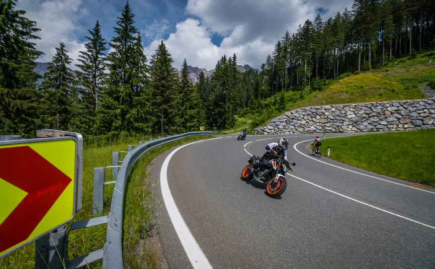 Motorradfahrer auf kurviger Bergstraße mit Fahrradfahrer und Wald