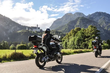 Two motorcyclists riding on mountain road with castle and mountains in background