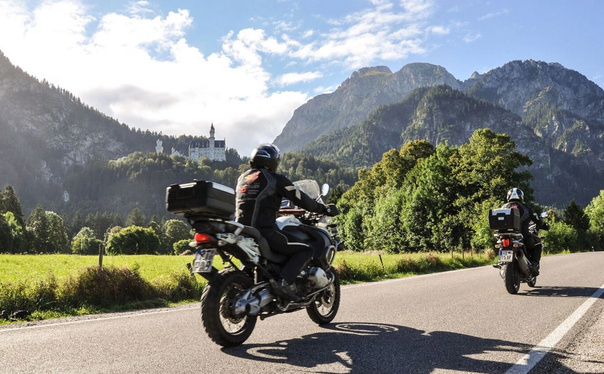 Zwei Motorradfahrer fahren auf einer Bergstraße mit Schloss und Bergen im Hintergrund