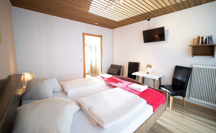 Helles Schlafzimmer mit Doppelbett, zwei Stühlen, Tisch und Fernseher an der Wand