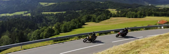 Zwei Motorradfahrer auf kurviger Bergstraße mit Bergen im Hintergrund