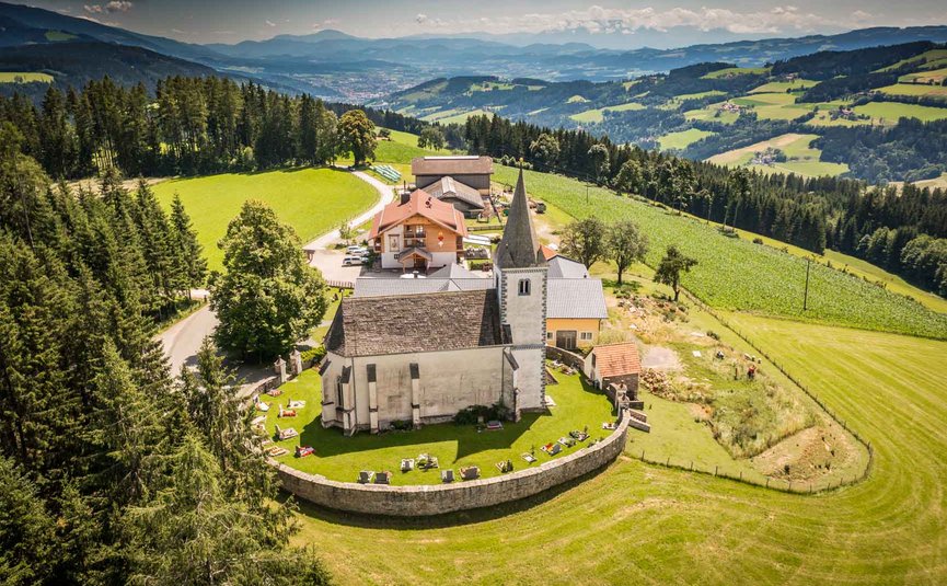 Kirche mit Friedhof auf grünem Hügel umgeben von Wäldern und Bergen