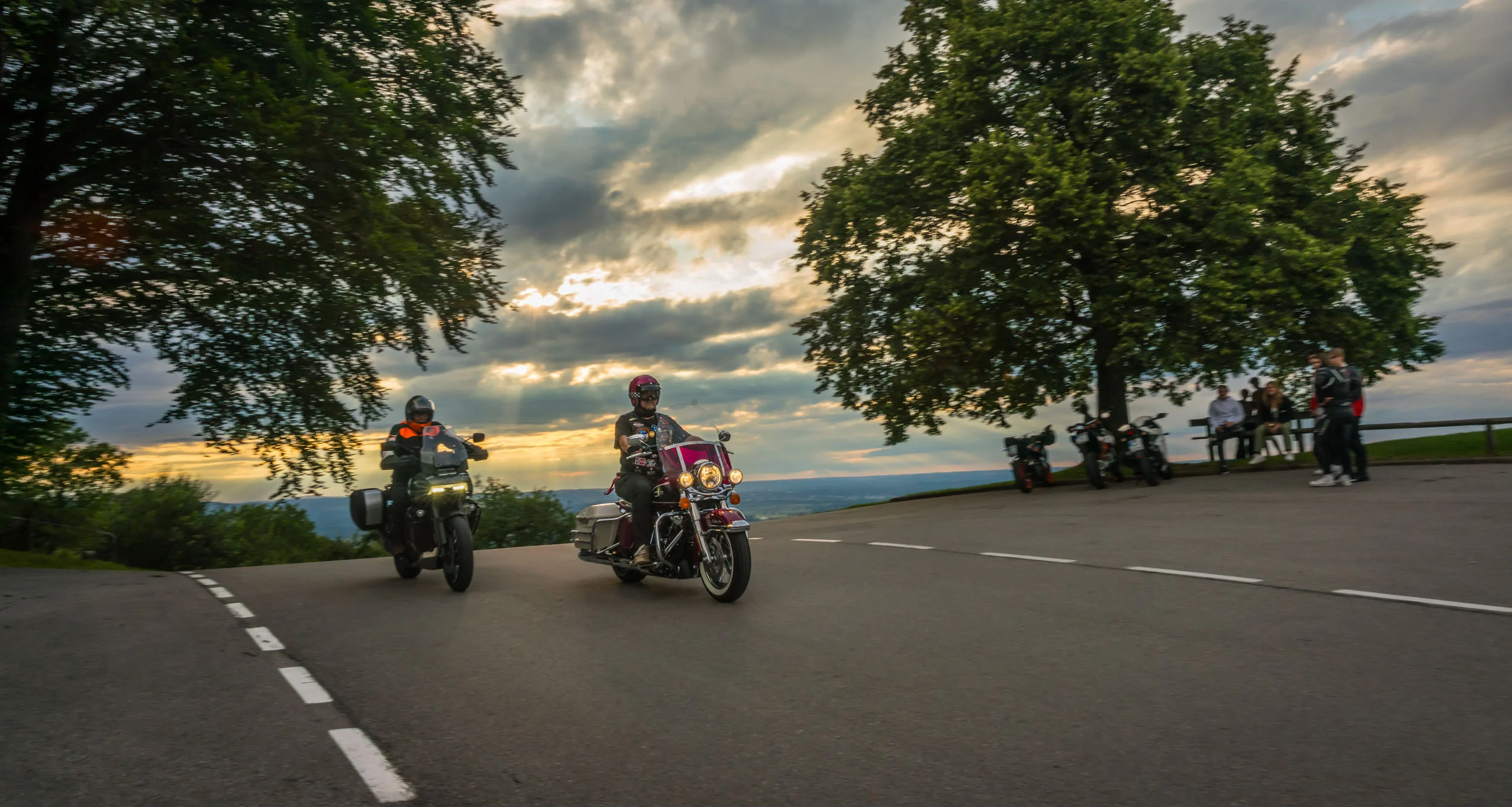 Zwei Motorradfahrer fahren bei Sonnenuntergang an einer Kurve vorbei.