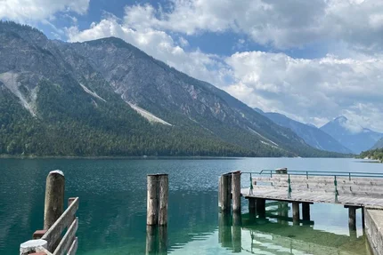 Wooden pier at clear mountain lake with forested mountains and cloudy sky