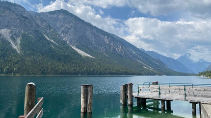 Holzsteg am klaren Bergsee mit bewaldeten Bergen und wolkigem Himmel