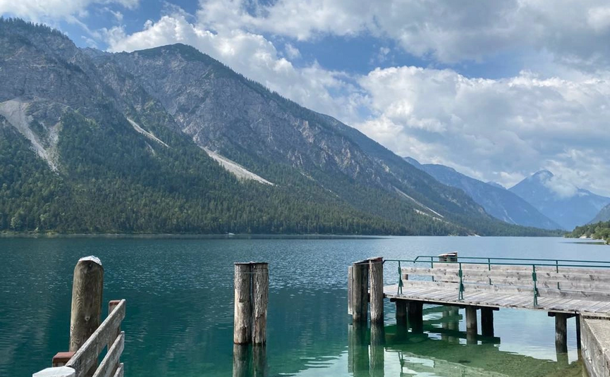 MoHo Schönauer Hof- Tour 9 Long Sleeper Variant 3 Wooden pier at clear mountain lake with forested mountains and cloudy sky
