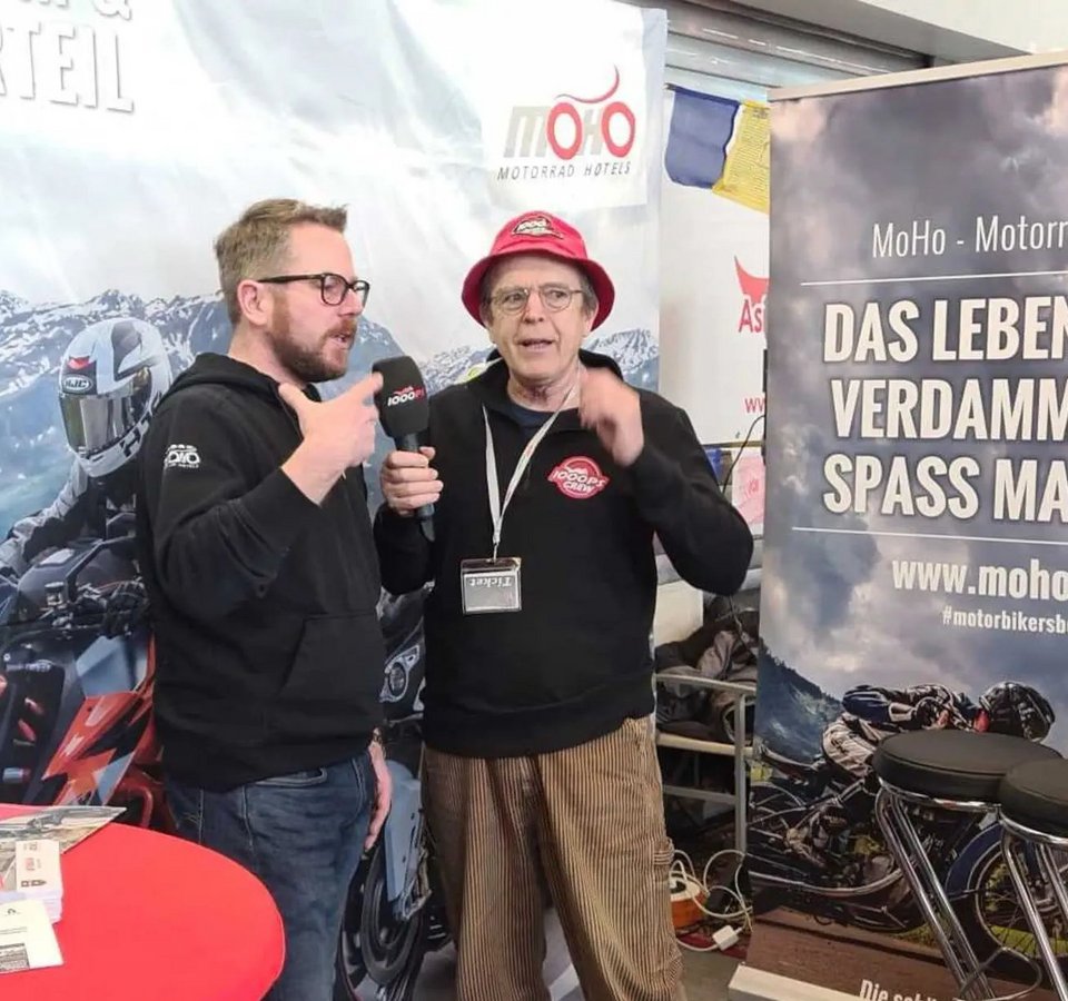 Moto Austria Zwei Männer im Gespräch auf einer Motorradmessen mit Werbebannern im Hintergrund