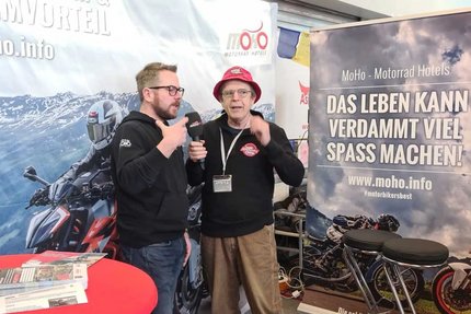 Zwei Männer im Gespräch auf einer Motorradmessen mit Werbebannern im Hintergrund