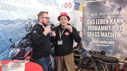Zwei Männer im Gespräch auf einer Motorradmessen mit Werbebannern im Hintergrund