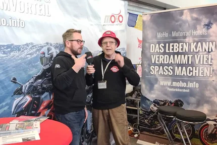 Twee mannen praten op een motorbeurs met reclamebanners op de achtergrond