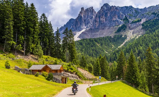 Motorcyclist riding on winding mountain road near wooden cabin and pine trees