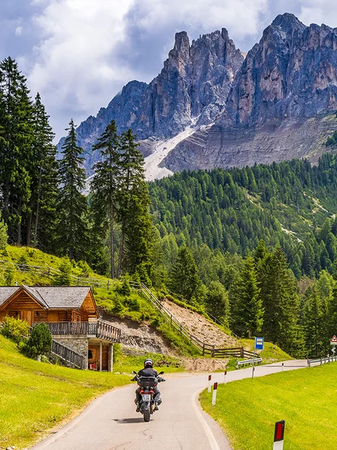 Motorcyclist riding on winding mountain road near wooden cabin and pine trees