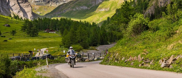 Motorradfahrer auf Bergstraße mit schneebedeckten Alpen und grüner Landschaft