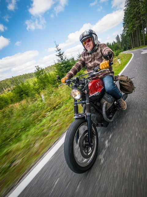 Older man riding motorcycle on winding country road through forest