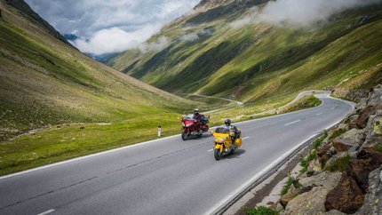 Arlberg – Dreiländereck © Moppetfoto.de Zwei Motorräder fahren auf einer Bergstraße mit grünen Hügeln und Wolken