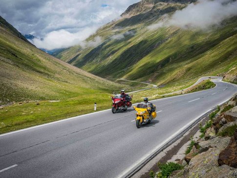 Two motorcycles riding on a mountain road with green hills and clouds