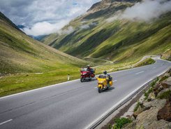 Arlberg – Dreiländereck © Moppetfoto.de Zwei Motorräder fahren auf einer Bergstraße mit grünen Hügeln und Wolken