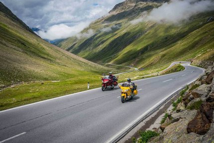 Zwei Motorräder fahren auf einer Bergstraße mit grünen Hügeln und Wolken