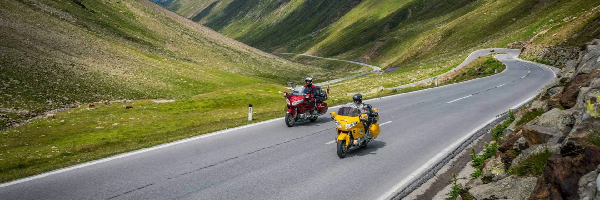 Two motorcycles riding on a mountain road with green hills and clouds