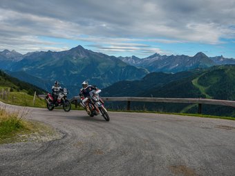 Zwei Motorradfahrer fahren auf Bergstraße mit Alpen im Hintergrund