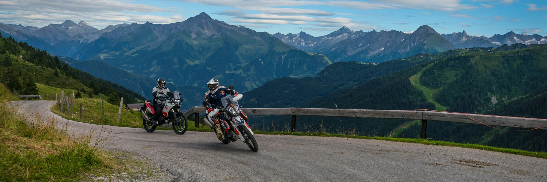 Two motorcyclists riding on a mountain road with Alps in the background