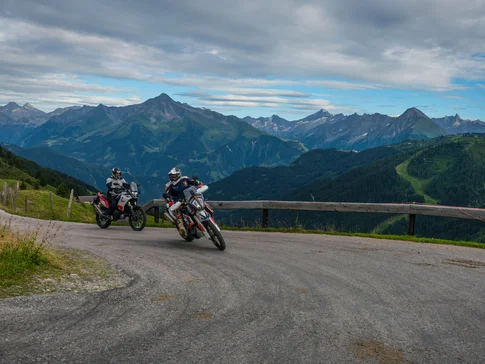 Two motorcyclists riding on a mountain road with Alps in the background