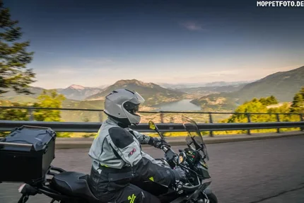 Motorradfahrer in Schutzkleidung fährt auf Bergstraße mit Berg- und Flussblick