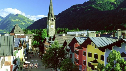 Kitzbüheler Hansi Tour (Gerlos - Zillertal - Kitzbühel) Bunte Häuserstraße und Kirchturm vor bewaldeten Bergen bei Sonnenschein