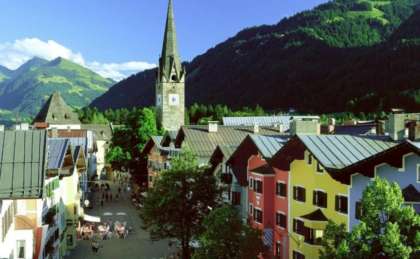 Kitzbüheler Hansi Tour (Gerlos - Zillertal - Kitzbühel) Bunte Häuserstraße und Kirchturm vor bewaldeten Bergen bei Sonnenschein